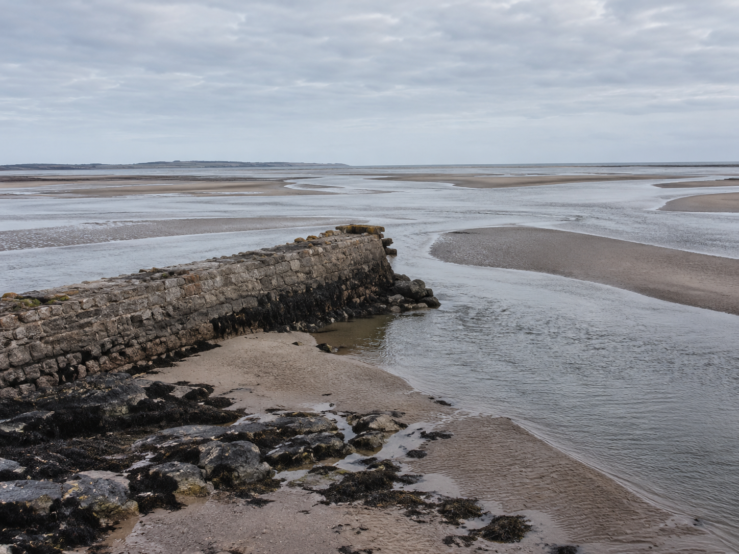 A weathered stone breakwater extends into a tidal estuary surrounded by pale sandbanks and branching shallow channels.